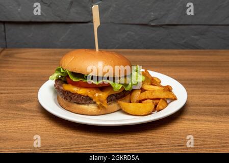 Klassischer Rindfleischburger mit geschmolzenem Cheddar-Käse, Tomaten, Eisbergsalat, eingelegten Gurken mit Keilfries Stockfoto