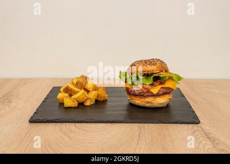 Klassischer Rindfleischburger, doppelt geschmolzener Cheddar-Käse, Zwiebeln und Tomaten, Mayonnaise und Ketchup, geräucherter Speck und Backkartoffeln Stockfoto