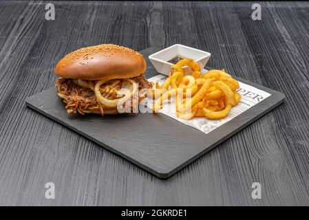 Pulled Pork Burger mit knusprigen weißen Zwiebelringen, Seed Bread, Curly Fries und BBQ Sauce Stockfoto