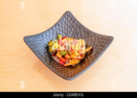 Riesengarnelen in Teriyaki-Sauce im Wok mit frischem Gemüse und Brokkoli mit Sesamsamen und Mohnblumen auf einer schwarzen Schüssel anbraten Stockfoto