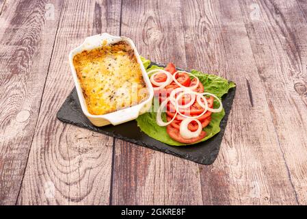 Einzelbehälter mit einer Portion Gratin Lasagne, begleitet von Tomaten- und Zwiebelsalat auf schwarzem Schiefer Stockfoto