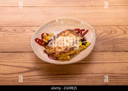 Beigefarbene Schüssel mit Gemüselasagne und Käse-Gratin mit Kürbisblüten und essbaren Blüten Stockfoto