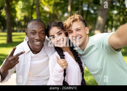 Multirassische Studenten befreunden sich, Selfie zu machen, V-Zeichen zu gestikulieren und Daumen hochzuzeigen, im Freien auf dem Campus zu posieren Stockfoto