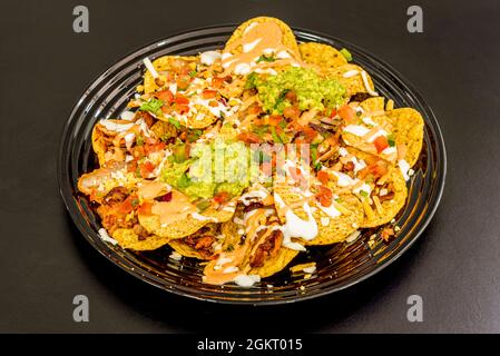 Teller mit runden Nachos mit zerfetztem Fleisch, Guacamole, Käse in der Menge mit Tomatenmark, Zwiebeln, Paprika und Petersilie auf schwarzem Teller Stockfoto