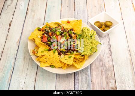 Vegane Nachos mit Jalapenos, Guacamole mit Petersilie und Schnittlauch und schwarze Bohnen mit Paprika, Tomaten und Zwiebeln auf Holztisch Stockfoto