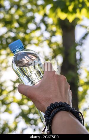 Weibliche Hand hält an einem heißen Sommertag eine Flasche mit kaltem Trinkwasser auf dem verschwommenen Hintergrund von Grün. Ansicht von unten. Stockfoto