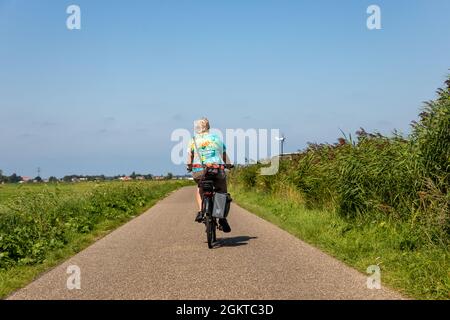 Ältere Dame, die auf einer asphaltierten Straße (Deich) mit einem Elektrofahrrad im südholländischen Dorf Lisse in den Niederlanden unterwegs ist. Von hinten aufgenommen. Mit einem Stockfoto