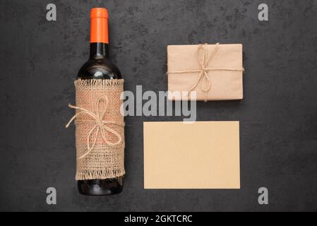 Rotwein-Flasche, Geschenkbox in Kraftpapier verpackt und leere Karte für Ihre Nachricht auf schwarzem Hintergrund. Null-Abfall-Urlaub, umweltfreundliche Geschenkverpackung Stockfoto