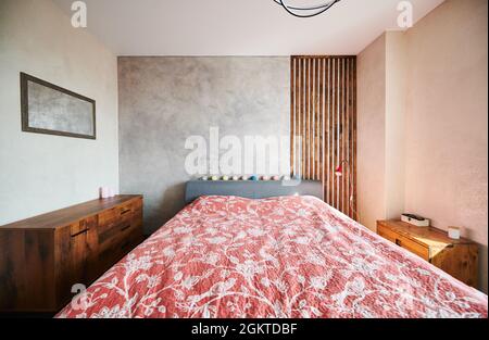 Blick auf das Design des Rutschzimmers mit großem Bett und roter Blumendecke. Schlafzimmer mit zwei hölzernen Kommode. Konzept der Schlafzimmer-Innenausstattung. Stockfoto