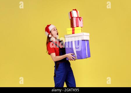 Aufgeregt Kurierin weiblich hält Stapel von Geschenken, Lieferung von Geschenken für Kunden für Winterferien, trägt blauen Overalls und weihnachtsmann Hut. Innen Stockfoto
