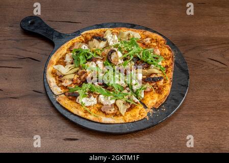 Kleine Pizza mit dünner Kruste mit Büffelkäse, Artischockenherzen, Rucola und Schweinestorozos auf einem schwarzen Tablett Stockfoto