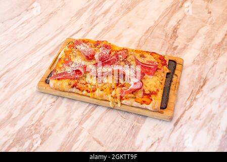 Rechteckige Pizza aus frischem Weizenteig mit serrano-Schinken und geriebenem Käse auf Marmortisch Stockfoto