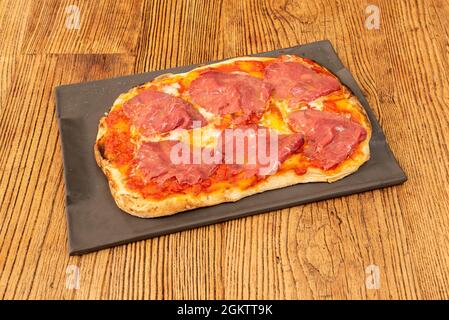 Rechteckige Pizza mit serrano-Schinken, Tomate und Mozzarella, die in einem Steinofen gebacken und auf einem schwarzen Teller serviert wird Stockfoto