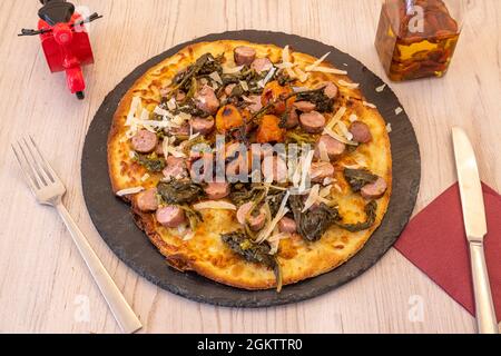 Pizza gefüllt bis zum Rand mit Weißwurst, Kirschtomaten, Rübengemüse und geriebenem Käse auf schwarzem Schiefer Stockfoto