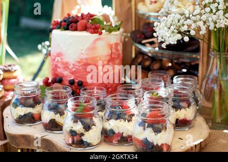 Cremiges Dessert mit frischen Beeren in einem Glas. Desserts stehen auf einem Holzblock, mit einem Geburtstagskuchen im Hintergrund. Fragment eines süßen Tisches Stockfoto