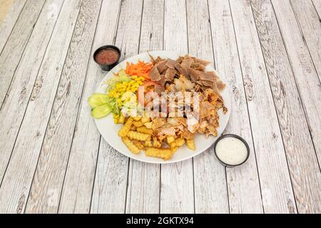 Gemischter Kebab-Teller mit Huhn und Lamm, garniert mit hausgemachten Pommes Frites, süßem Mais, geriebener Karotte, Gurkenscheiben und Saucen auf weißem Tisch Stockfoto