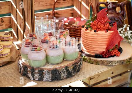 Cremiges Dessert mit frischen Beeren in einem Glas. Desserts stehen auf einem Holzblock, mit einem Geburtstagskuchen im Hintergrund. Fragment eines süßen Tisches Stockfoto