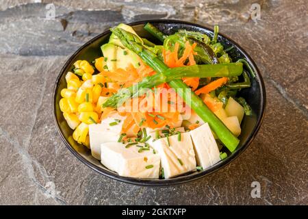 Vegane Schüssel mit wildem Spargel, Karotte, Tofu-Stücke, zerkleinerten Schnittlauch, süßem Mais, Avocado, Wakame-Sauce und Ananas auf grauem Stein Stockfoto