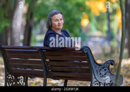 Ältere Frau, die im Herbstpark auf der Bank sitzt und die Kamera anschaut. Stockfoto