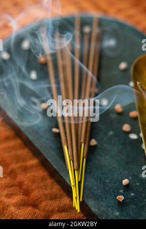 Selektiver Fokus von Weihrauch auf einer Steintafel und Räucherstäbchen auf der Seite, orangefarbenem Stoffhintergrund und Rauch im Vordergrund Stockfoto