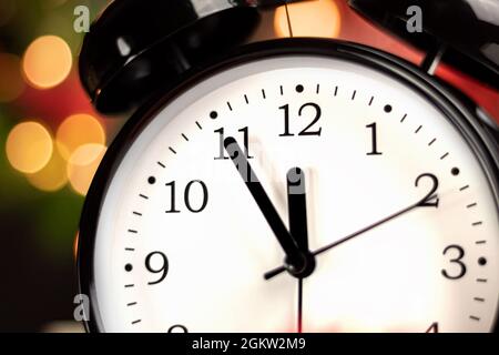 Eine Uhr in Neujahrsdekorationen ist mit Fichtenzweigen und bunten Lichtern umhüllt. Die Uhr zeigt die Zeit an: Fünf Minuten bis Mitternacht. Hintergrund w Stockfoto