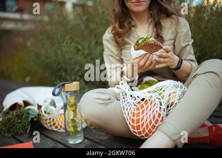 Nahaufnahme der Frau im Ganzen mit Saitenbeutel, Wasserflasche und Sandwich beim Essen im Freien in der Stadt. Stockfoto