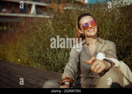 Lächelnde, elegante Frau mittleren Alters in Sonnenbrillen und insgesamt mit einer Tasche, die draußen in der Stadt sitzt. Stockfoto