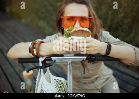 Trendige 40 Jahre alte Frau in Sonnenbrille und insgesamt mit Saitentasche, Roller und Sandwich draußen in der Stadt sitzen. Stockfoto
