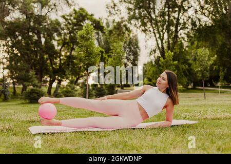 Attraktive, elegante Frau ist in Pilates mit Ball auf grünem Gras des Rasens bei Sonnenuntergang beschäftigt Stockfoto