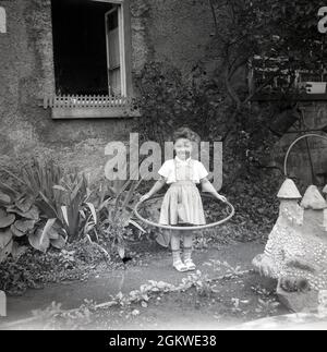 1960er, historisch, draußen auf einem Gartenweg, ein kleines Mädchen mit einem Hula Hoop Reifen, einem Spielzeugreifen, um ihre Taille, Deutschland. Obwohl es den Hula Hoop Reifen schon seit Hunderten von Jahren gab und von Kindern benutzt wurde, begann erst in den späten 1950er Jahren, als die Plastikversion eingeführt wurde, der Wahns für sie. Der Hula Hoop Reifen wurde zu einer weltweiten Modeerscheinung, mit mehr als 100 Millionen verkauften in zwei Jahren. Hula Hoops blieb bis in die späten 1970er, frühen 80er Jahre ein beliebtes Kinderspielzeug. Stockfoto