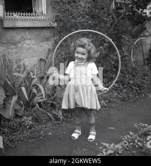 1960er, historisch, draußen auf einem Gartenweg, ein kleines Mädchen mit einem Hula Hoop Reifen, einem Spielzeugreifen, um ihre Taille, Deutschland. Obwohl es den Hula Hoop Reifen schon seit Hunderten von Jahren gab und von Kindern benutzt wurde, begann erst in den späten 1950er Jahren, als die Plastikversion eingeführt wurde, der Wahns für sie. Der Hula Hoop Reifen wurde zu einer weltweiten Modeerscheinung, mit mehr als 100 Millionen verkauften in zwei Jahren. Hula Hoops blieb bis in die späten 1970er, frühen 80er Jahre ein beliebtes Kinderspielzeug. Stockfoto