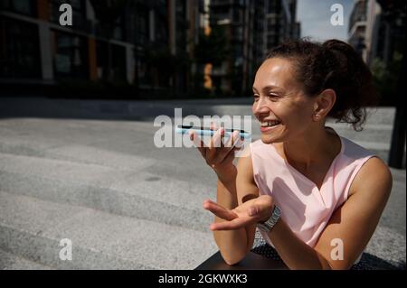Liebenswert gemischte Rennen schöne Frau Aufnahme einer Sprachnachricht auf dem Smartphone, ruht auf Stufen auf dem Hintergrund des Gebäudes Stockfoto