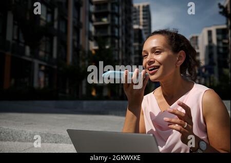 Liebenswert gemischtes Rennen schöne Frau Aufnahme einer Sprachnachricht auf dem Smartphone während der Arbeit auf Laptop sitzen auf Stufen auf dem Gebäude Hintergrund. Busi Stockfoto