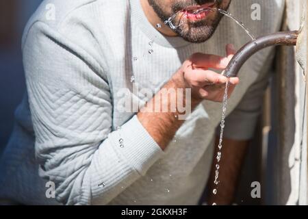 Der kaukasische Mann trinkt Wasser aus einem historischen traditionellen Trinkbrunnen im Freien in Rom. Heißer Sommertag. Lässiger weißer Pullover, blaue Jeans Stockfoto