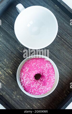 Vertikales Bild von leuchtend rosa Donut auf weißem runden Teller und Teetasse oben. Alter Holzhintergrund. Isolierte Objekte. Traditionelles amerikanisches Frühstück Stockfoto