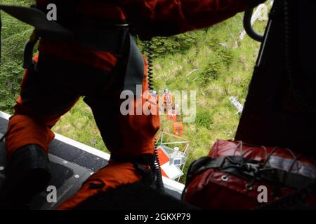 Ein Mitglied der Küstenwache der Flugstation Sitka beobachtet zwei Küstenwache-Aids für das Navigations-Team Sitka-Mitarbeiter, die Wartungsarbeiten an ATON in Sitka, Alaska, durchführen, 21. Juli 2021. Wenn Besatzungen auf ATON in abgelegenen Gebieten Alaskas arbeiten, werden Hubschrauber häufig eingesetzt, um die Besatzung und Ausrüstung dorthin zu bringen. Stockfoto