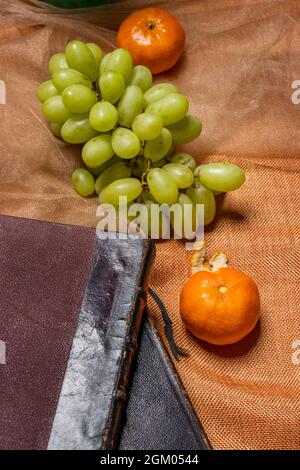 Ein Bund länglicher weißer Desserttrauben mit reifen Mandarinen auf orangefarbenem Tuch und alten Büchern Stockfoto