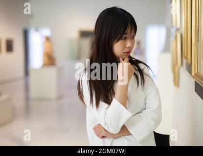 Nachdenkliche chinesische Frau, die im Kunstmuseum in der Nähe des Gemäldes steht Stockfoto