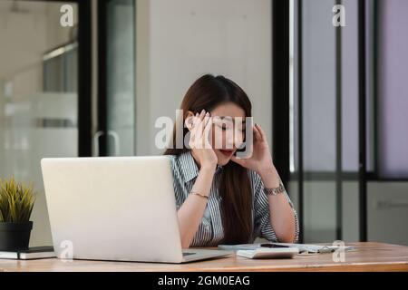 Gestresste junge Frau, die am Schreibtisch sitzt und vom Heimbüro aus arbeitet, Geschäftsfrau Kopfschmerzen, während sie ein Problem bei der Arbeit im Büro hat Stockfoto