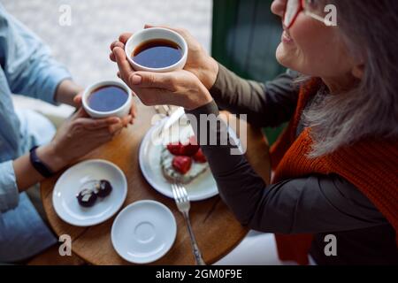 Lächelnde reife Frau mit Freund halten Tassen Tee an einem kleinen Tisch im Café Stockfoto