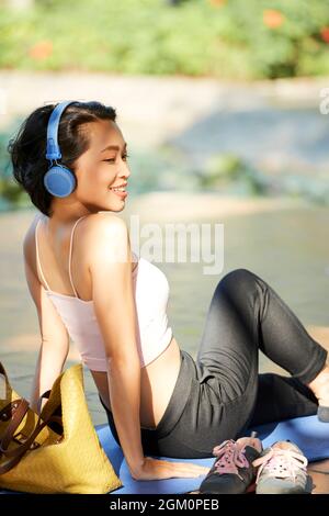 Fröhliche, positive junge Frau, die sich draußen auf einer Yogamatte ausruhen und Musik über Kopfhörer hören kann Stockfoto