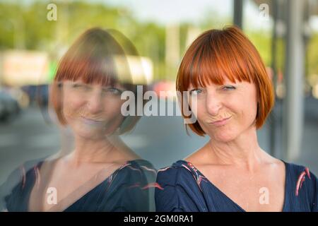 Dubiose Rothaarige Frau, die der Kamera einen skeptischen, ungläubigen Blick verleiht, als sie sich an ein großes Schaufenster lehnt, das sich im Glas widerspiegelt Stockfoto