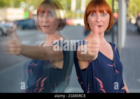 Eine attraktive, enthusiastische Rothaarige Frau, die mit einem triumphorischen Lächeln einen Daumen nach oben drückt, während sie sich an ein Schaufenster lehnt, das sich im Glas spiegelt Stockfoto