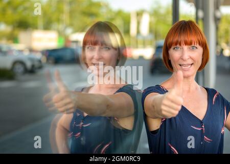 Eine attraktive, enthusiastische Rothaarige Frau, die mit einem triumphorischen Lächeln einen Daumen nach oben drückt, während sie sich an ein Schaufenster lehnt, das sich im Glas spiegelt Stockfoto