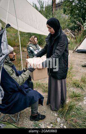 Muslimische Frau mit schwarzem Kopftuch und großer Jacke, die sich im Flüchtlingslager mit jungen Flüchtlingen austeilt Stockfoto