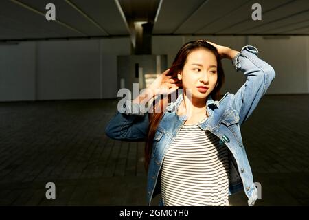 Porträt einer schönen jungen Frau in einer Jeansjacke, die ihre Haare berührt und wegschaut Stockfoto