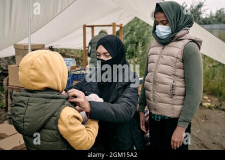 Muslimische Frau in Hijab, die ihre junge Tochter im Flüchtlingszelt mit Stoffen und Lebensmitteln kleidet Stockfoto