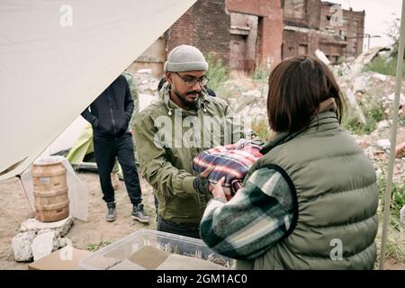 Rückansicht eines Sozialarbeiters in warmer Weste, der im Freien in Brille und Hut einem Migranten aus dem Nahen Osten karierten Stockfoto