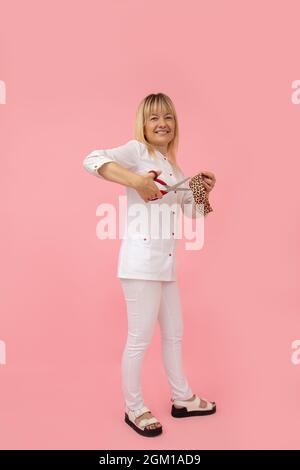 Portrait des blonden Kosmetologen in weißer medizinischer Uniform, zeigt, wie man Bandstreifen für das Gesichts-Taping schneidet. Anheben der Stirnseite. Schönheitssalons. Beauty .Mode Stockfoto