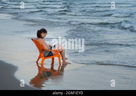 Tel Aviv, Israel - 20. August 2021: Eine Frau, die im Morgengrauen am Strand von Tel Aviv, Israel, auf einem orangefarbenen Strandstuhl sitzt und ihr Smartphone benutzt. Stockfoto